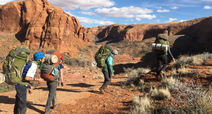 A group of people wearing backpacks hike along a trail through a desert landscape. There are tall canyon walls in the background.
